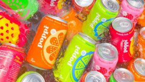 Latas de refrigerante prebiótico Poppi em diferentes sabores mergulhadas em gelo, destacando a recente aquisição da marca pela PepsiCo.