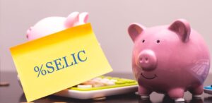 A imagem mostra um cofre de porquinho com moedas em volta e um papel escrito Selic para representar a taxa Selic.