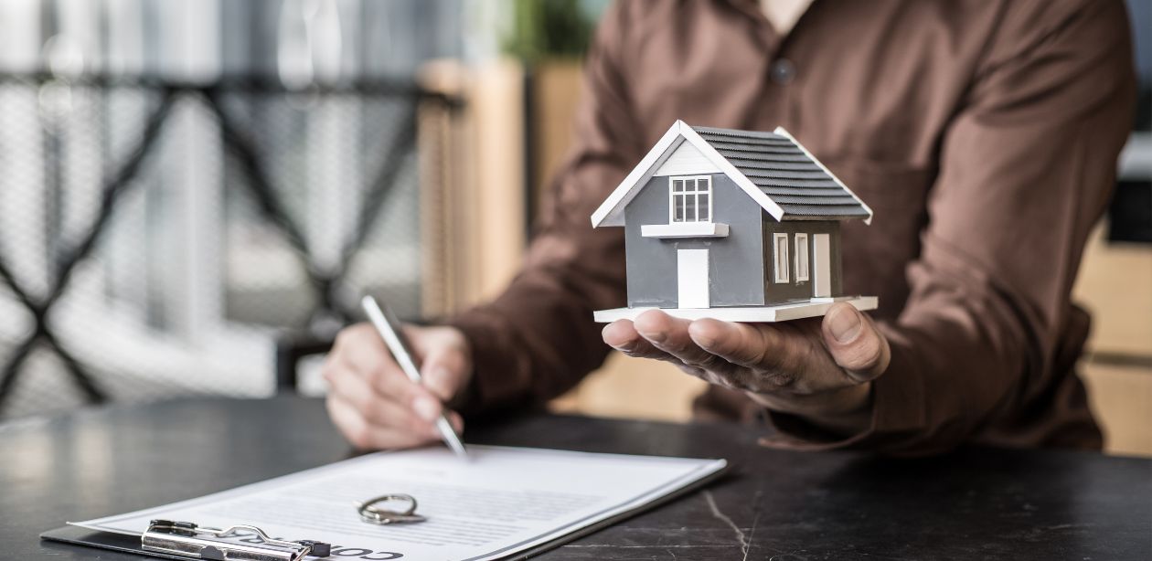 A imagem mostra uma pessoa segurando uma miniatura de uma casa e assinando um papel para representar que a venda de imóveis disparou.