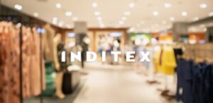 A imagem mostra o interior de uma loja de roupas ao fundo desfocada e a logo da Inditex destacada no centro da imagem para representar que as vendas da Inditex caem.
