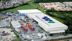 Vista aérea do centro logístico da Wilson Sons, com galpões, contêineres e caminhões em operação, destacando a marca no telhado.