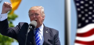 Donald Trump discursa com o punho erguido em um evento ao ar livre, enquanto a bandeira dos Estados Unidos tremula ao fundo.