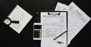 A imagem mostra vários papéis, calculadora e caneta sob uma mesa para representar a restituição do IRPF 2025