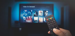 A imagem mostra uma pessoa segurando um controle remoto e ao fundo uma TV para representar que a Strima fortalece setor de streaming.