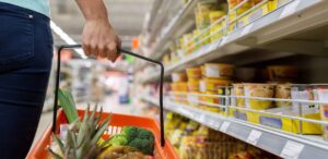 Pessoa segurando cesta de compras com vegetais, observando prateleiras diante do aumento no custo da cesta básica.