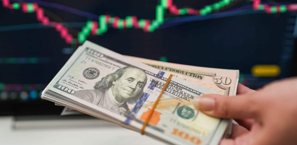 Maço de cédulas de dólar americano com gráficos do mercado financeiro, refletindo a cotação do dólar hoje.