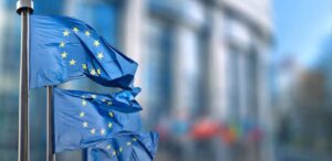 Bandeiras da União Europeia tremulando ao vento em frente a um prédio institucional.