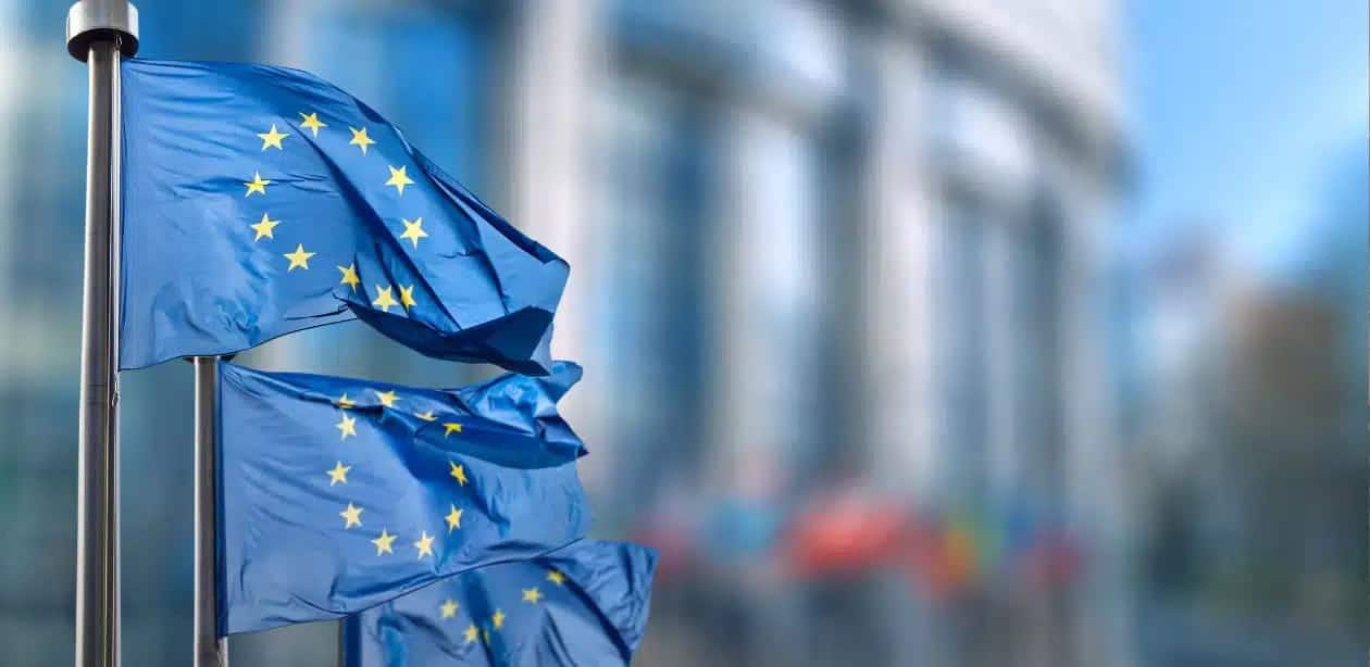 Bandeiras da União Europeia tremulando ao vento em frente a um prédio institucional.