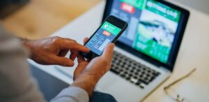 Pessoa utilizando smartphone para acompanhar apostas online no Brasil, com computador exibindo plataforma de apostas esportivas ao fundo.