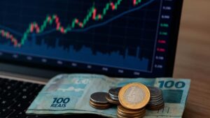 Notas de 100 reais e moedas em frente a gráfico de ações, representando juros sobre capital próprio do Banco Pine