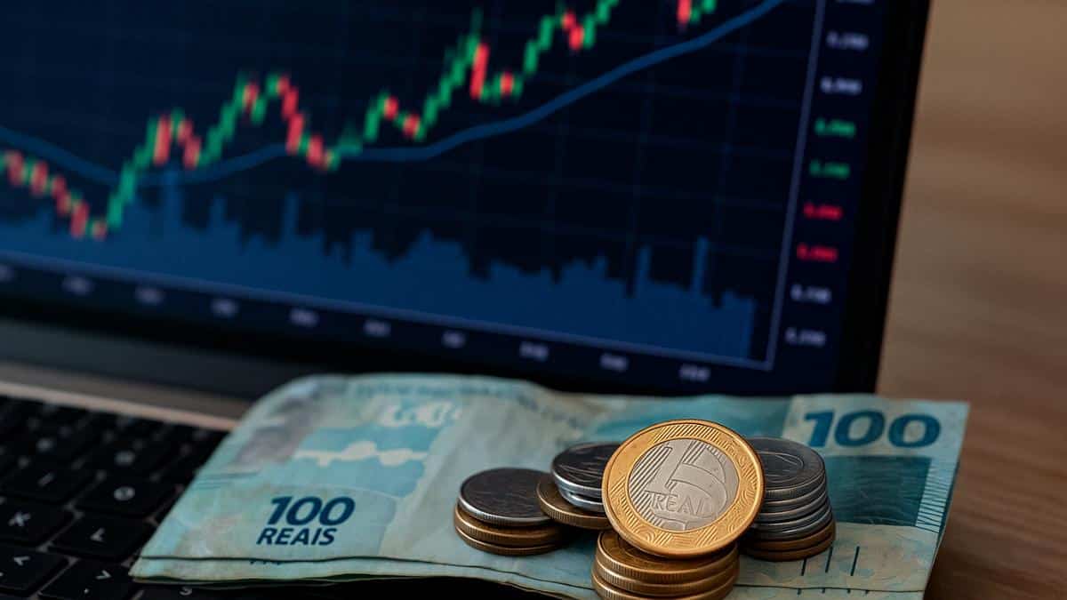 Notas de 100 reais e moedas em frente a gráfico de ações, representando juros sobre capital próprio do Banco Pine