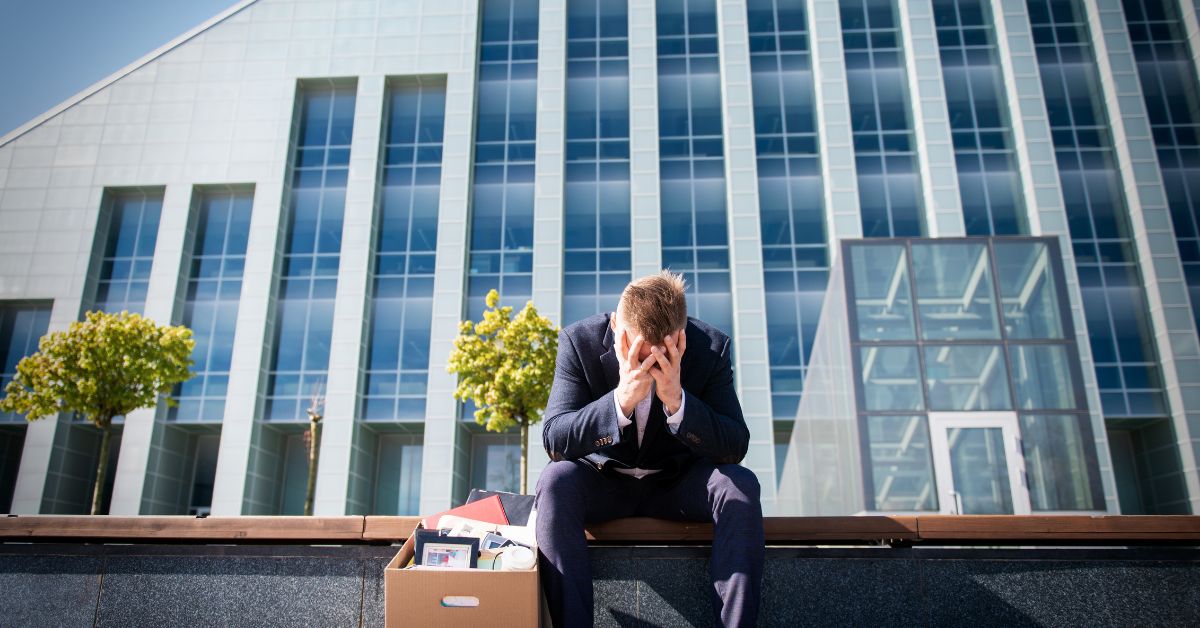 A imagem mostra um homem sentado de fora de uma empresa com seus pertences para representar que o desemprego sobe em 12 estados