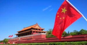 A imagem mostra a bandeira da China na frente de um monumento para representar que as ações da China disparam com trégua tarifária com os EUA