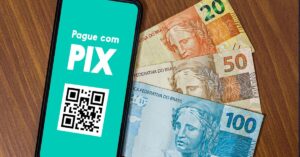 A imagem mostra um qr code de pix na tela de um celular ao lado de notas de dinheiro para representar a Plataforma de criptomoedas Binance