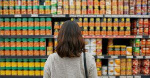 A imagem mostra uma mulher olhando preços em um supermercado para representar que o Índice de Preços ao Produtor (IPP) recua novamente 0,62% em março