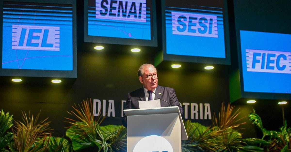 Presidente da FIEC discursa em cerimônia do Dia da Indústria, com painéis do IEL, SENAI, SESI e FIEC ao fundo do palco