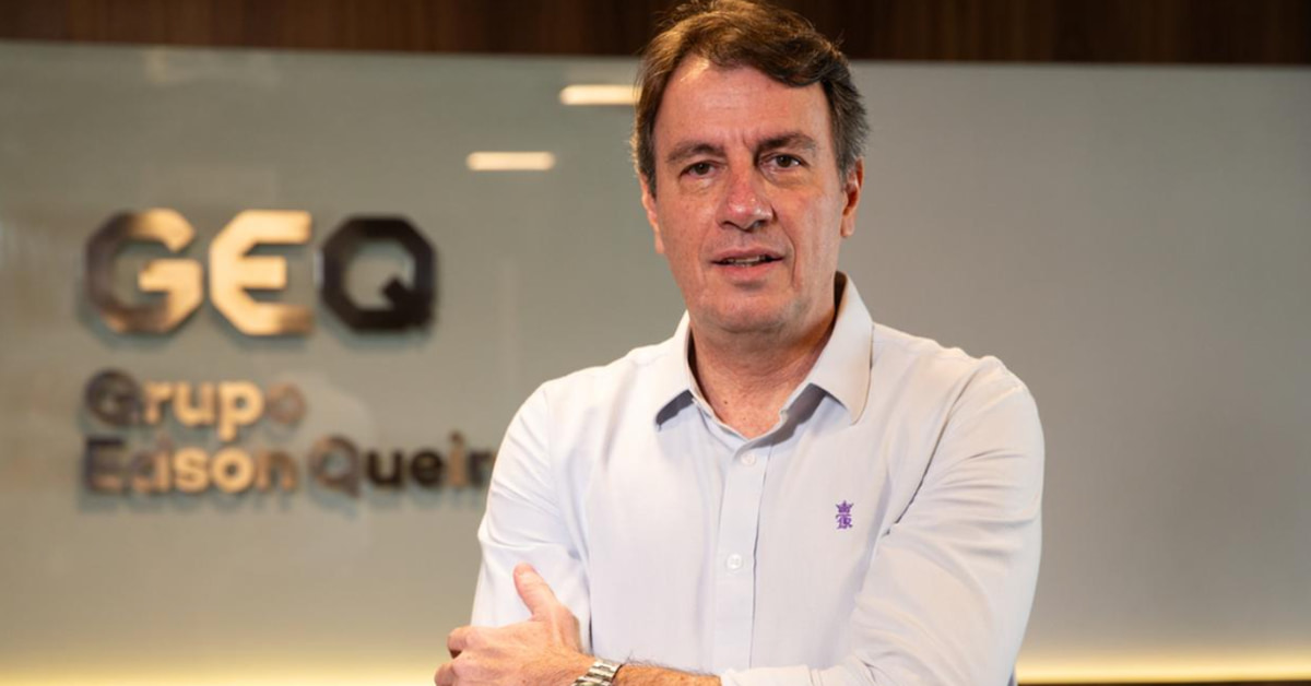 Carlos Rotella, presidente-executivo do Grupo Edson Queiroz (GEQ), aparece em pé com os braços cruzados, vestindo camisa social branca com um discreto símbolo roxo. Ao fundo, a logomarca da empresa está parcialmente visível em uma parede de vidro.