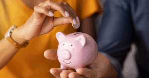 Mão colocando moeda em cofre rosa de porquinho, ilustrando o saldo da poupança em maio de 2025.