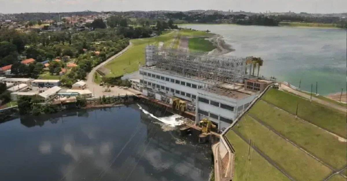 Aérea de usina hidrelétrica com estrutura de subestação sobre o prédio, vertedouro em operação e reservatório ao fundo.