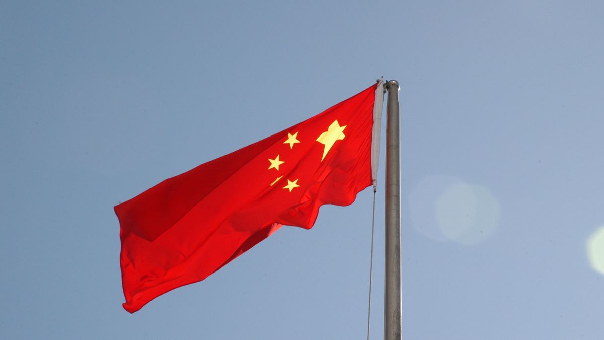 imagem ilustrativa mostra a bandeira vermelha da China, com cinco estrelas douradas, hasteada em um mastro metálico sob céu claro, simbolizando o país no contexto da crise das terras-raras.
