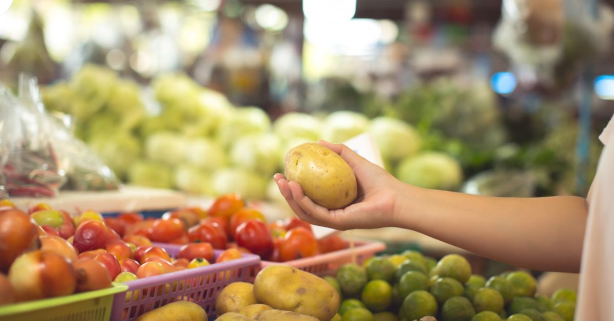 Pessoa segurando batata em banca de feira com frutas e verduras ao fundo