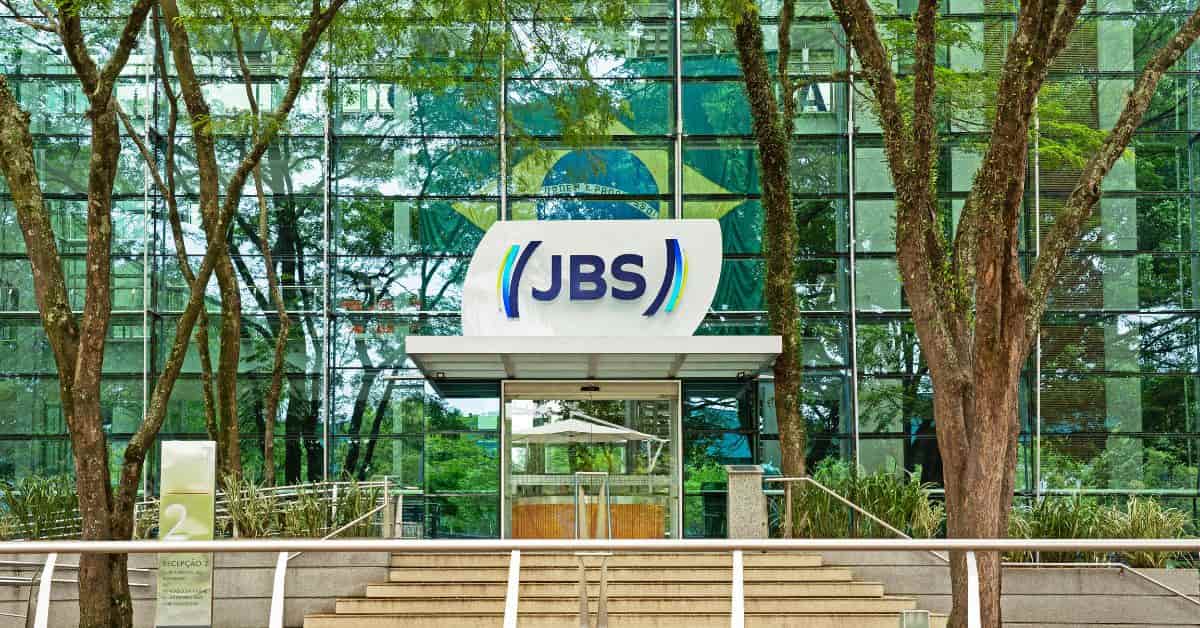 Fachada da sede da JBS com logotipo da empresa em destaque na entrada de edifício corporativo cercado por árvores.