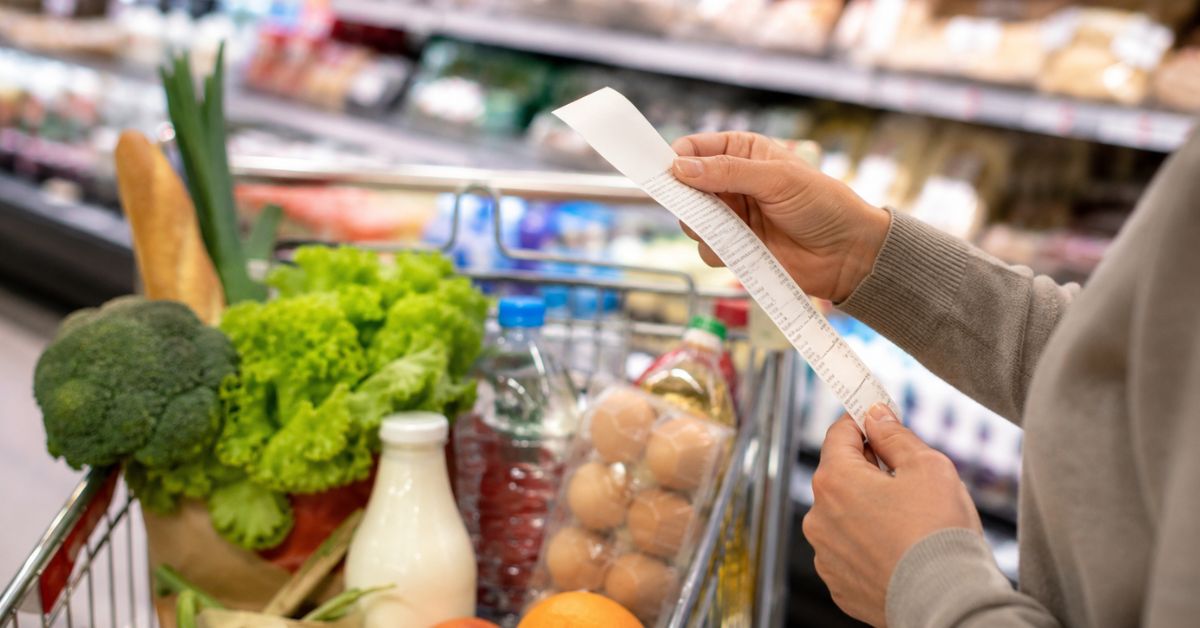 Pessoa segura recibo de compras em supermercado enquanto carrinho com alimentos básicos aparece em primeiro plano.