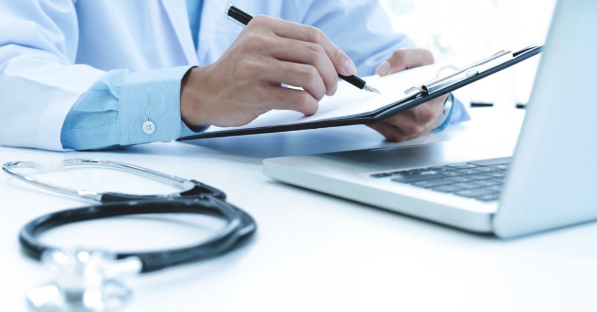 Médico preenche documentos ao lado de computador durante atividade administrativa relacionada à gestão de planos de saúde.
