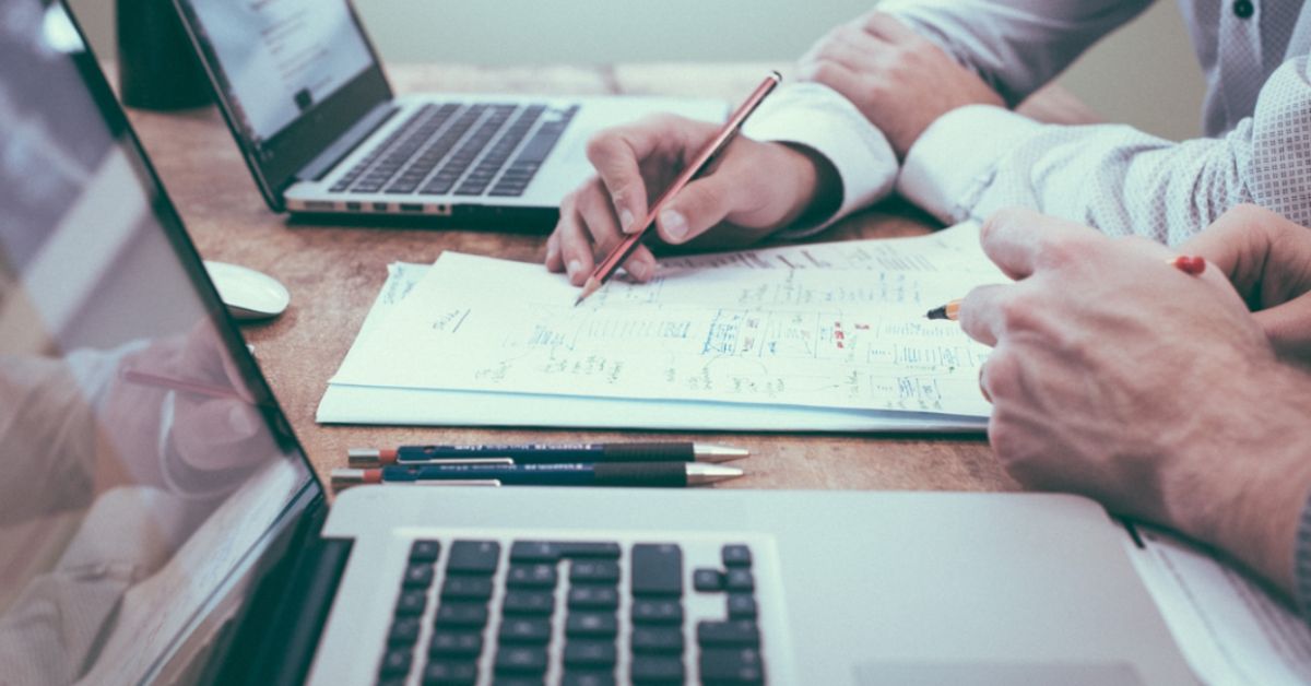 Profissionais analisam documentos e indicadores financeiros em mesa de reunião com notebooks e relatórios impressos.