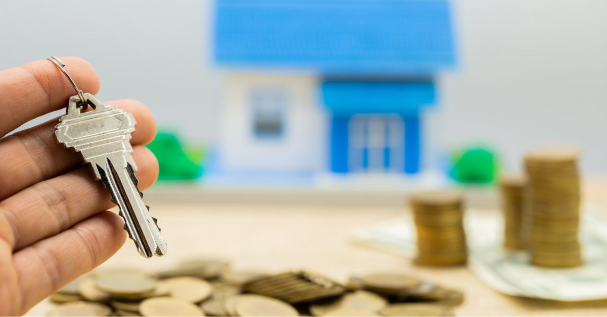 Mão segura chave de imóvel em primeiro plano enquanto miniatura de casa e moedas aparecem ao fundo, simbolizando o mercado de aluguel residencial.