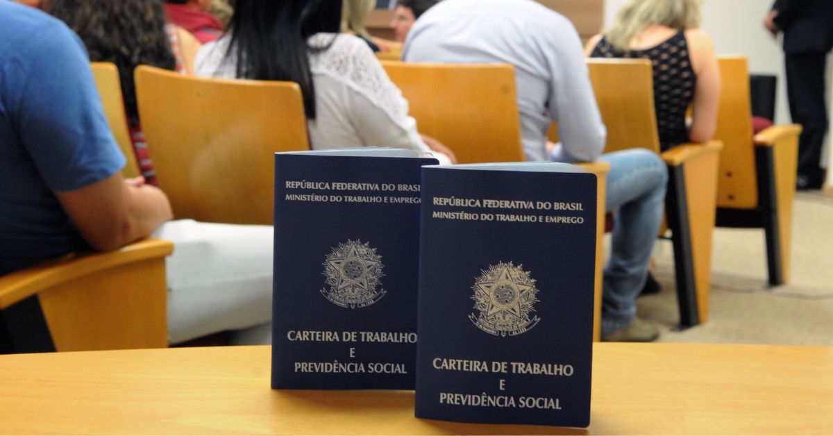 Carteiras de trabalho sobre mesa durante atendimento em unidade de emprego, com trabalhadores aguardando em cadeiras ao fundo.