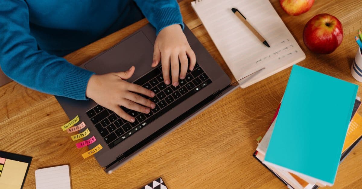 Pessoa utiliza notebook em mesa de trabalho enquanto consulta anotações e materiais de estudo, em ambiente associado à produção de conteúdo e análise de informações financeiras.