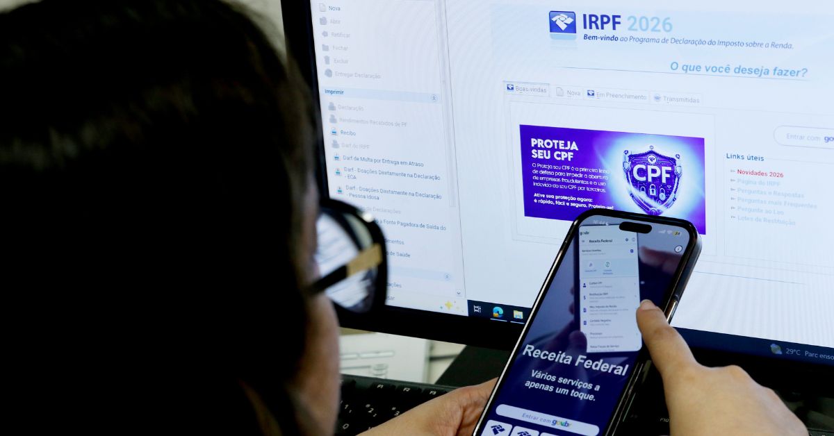 Pessoa utiliza o aplicativo da Receita Federal no celular enquanto acessa o programa do Imposto de Renda 2026 em um computador.