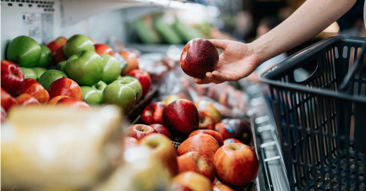 Consumidora escolhe maçãs em seção de frutas de supermercado durante compras, em imagem que representa o consumo das famílias e o desempenho do varejo.